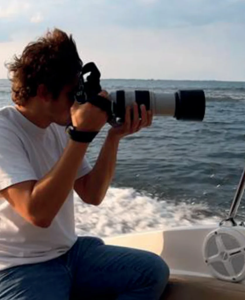 Person on a boat taking a photo with a large telephoto camera lens toward the ocean, waves in the background.
