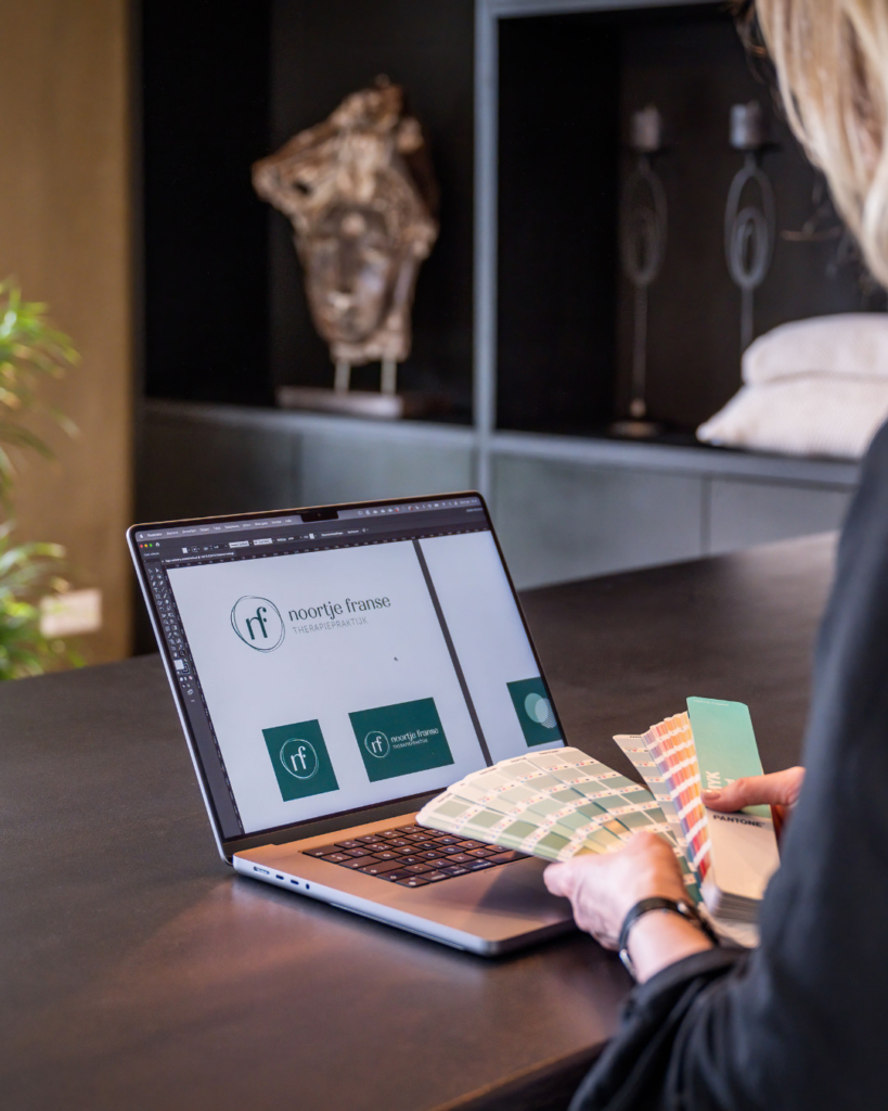 Person holds color swatches while working on a laptop displaying a branding mockup on screen site logo visible in design software