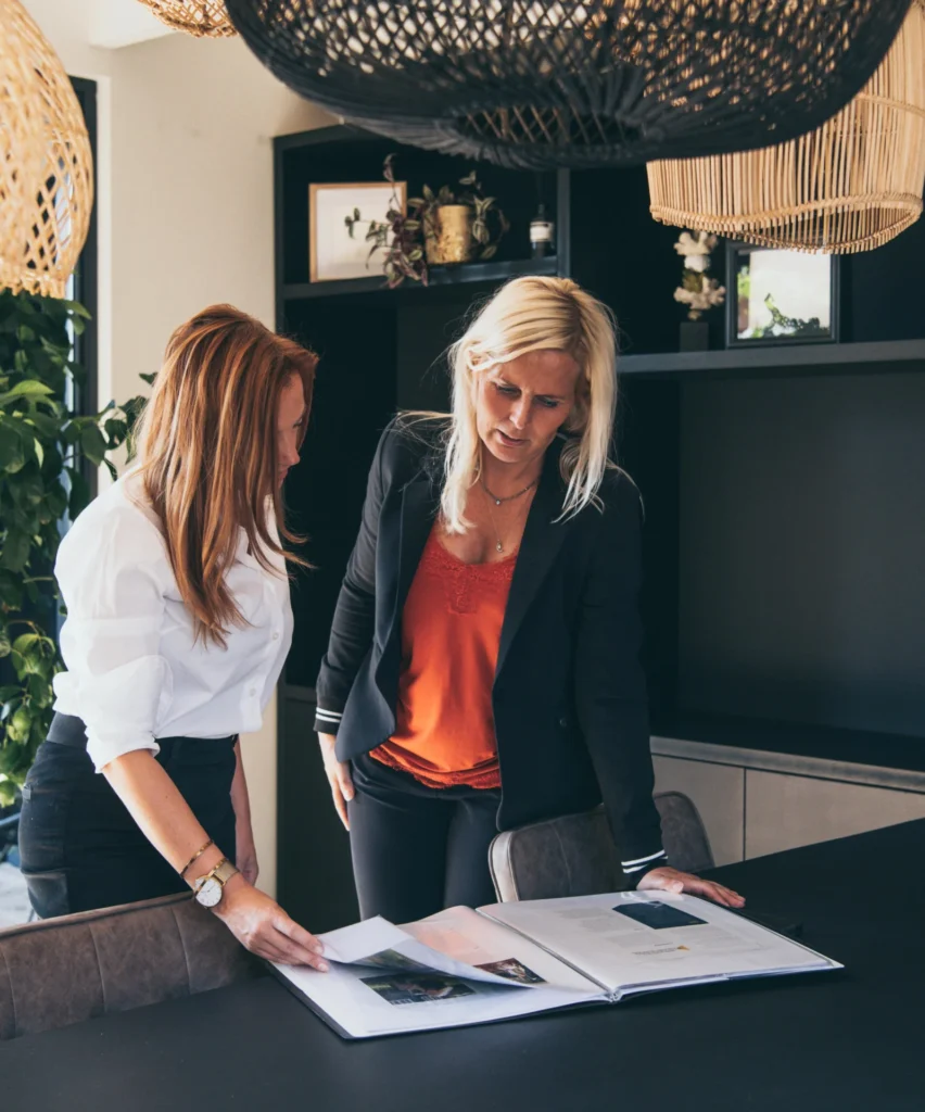 Two women review a large photo book spread open on a dark table in a stylish office setting.