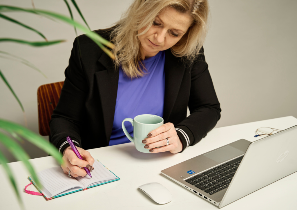 Woman at a white desk writing in a colorful notebook with a purple pen, holding a light blue mug; laptop and glasses nearby.