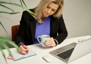 Woman in a blazer writes in an open notebook at a white desk, holding a mint mug with a laptop and glasses nearby.
