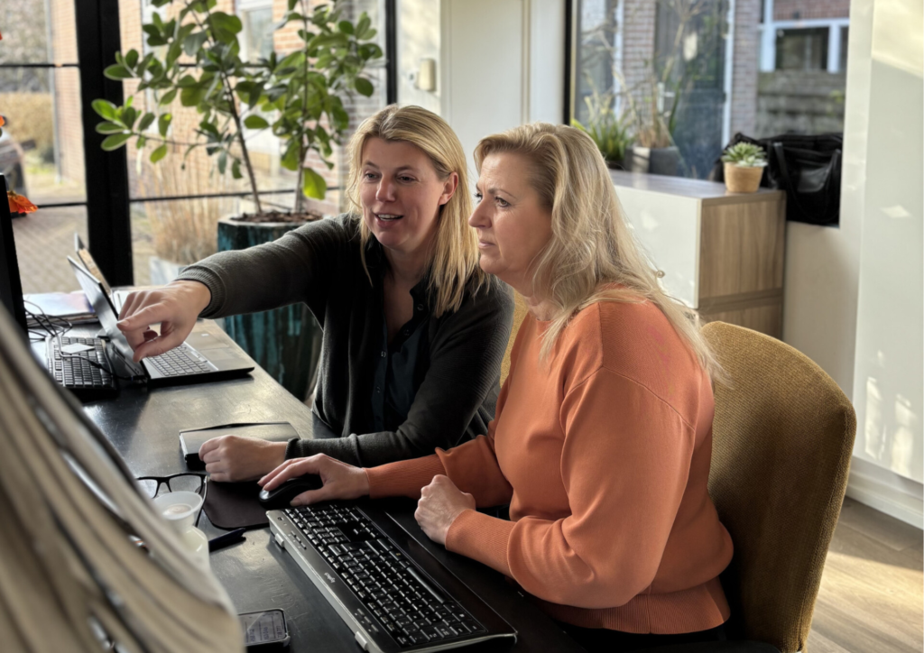 Two women collaborate at a desk, one pointing at the laptop screen as the other watches nearby in a bright office.