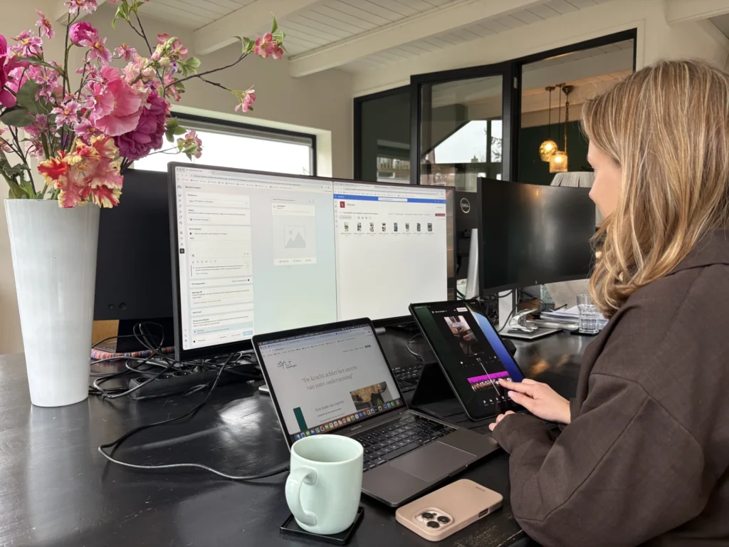Person at a workspace with two computer monitors, a laptop, a tablet, a mug, and a phone on a dark desk near a vase of pink flowers.
