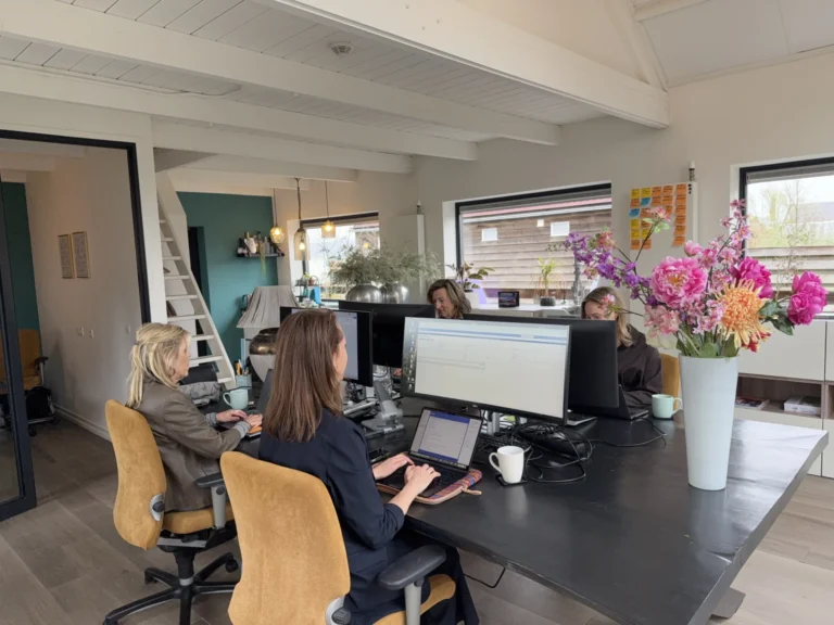 Open-plan office with four women at a large shared desk, multiple monitors, and a tall vase of pink flowers.