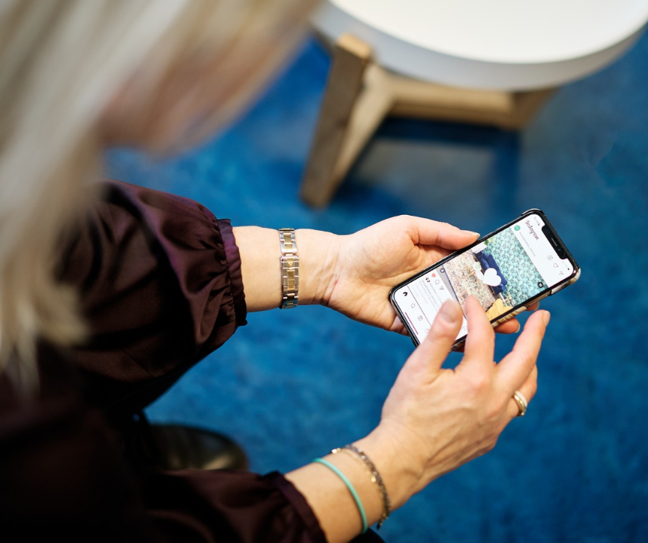 Close-up of a person’s hands holding a smartphone, scrolling/tapping the screen, wearing a silver watch and bracelets on a blue-carpeted surface.