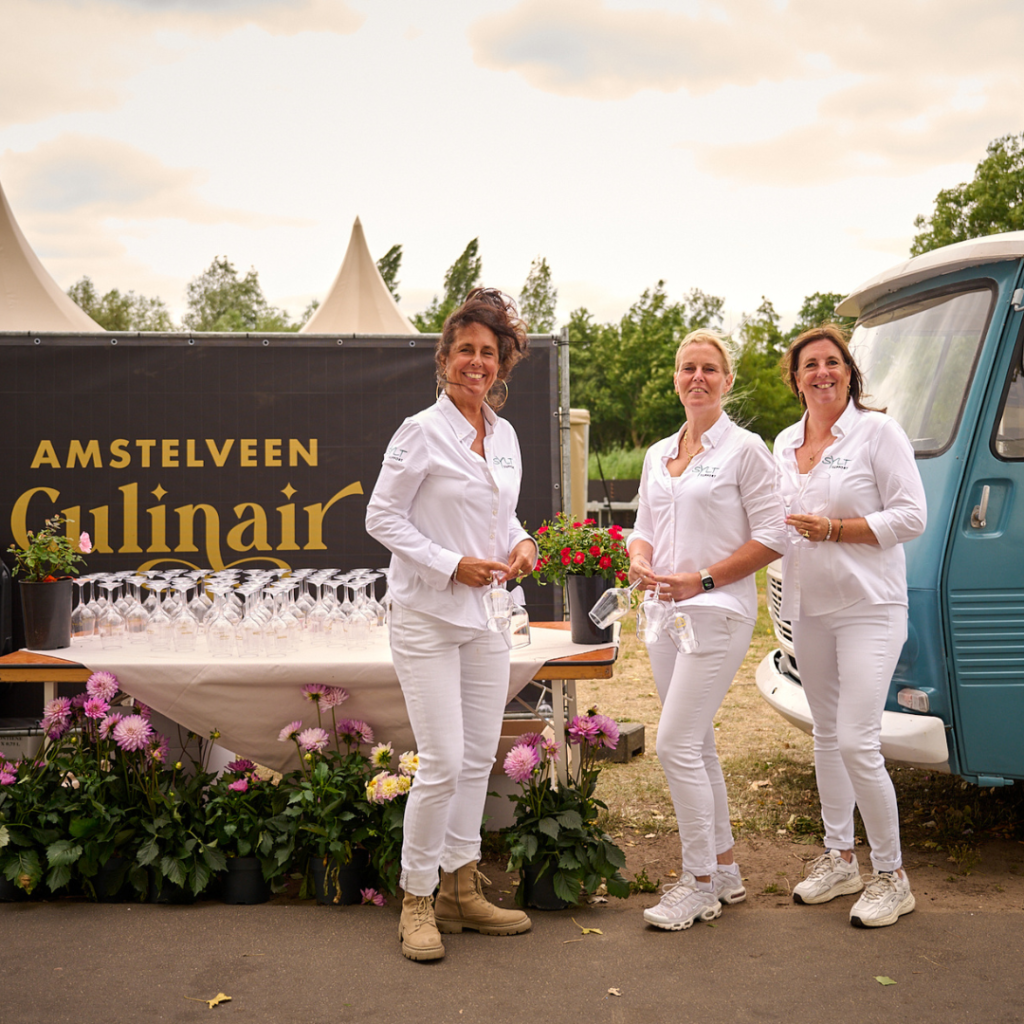 Three women in white outfits holding wine glasses at an outdoor event beside a blue vintage truck and a table of glasses; banner reads Amstelveen Culinair.