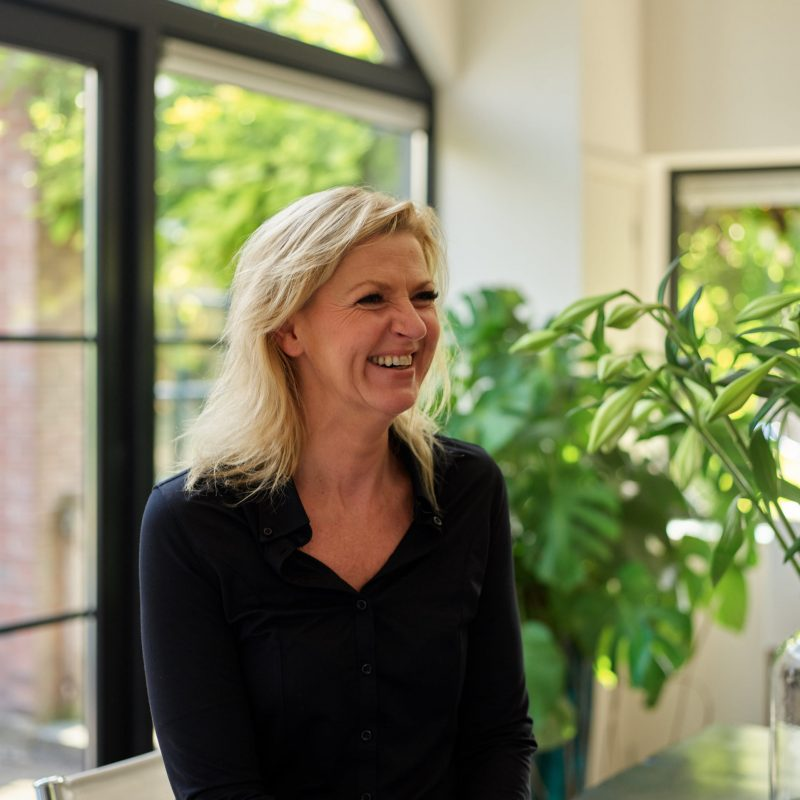 Smiling woman in a black button-down shirt indoors, near a large leafy plant by a windowed wall.