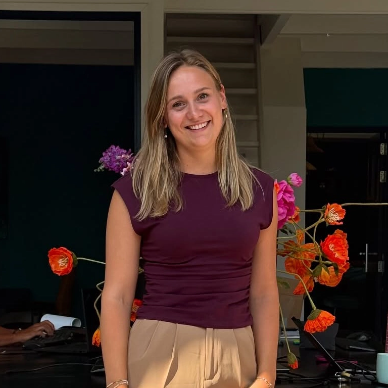 Smiling woman with blonde hair wearing a burgundy top and beige pants, standing indoors with bright orange flowers in the background.