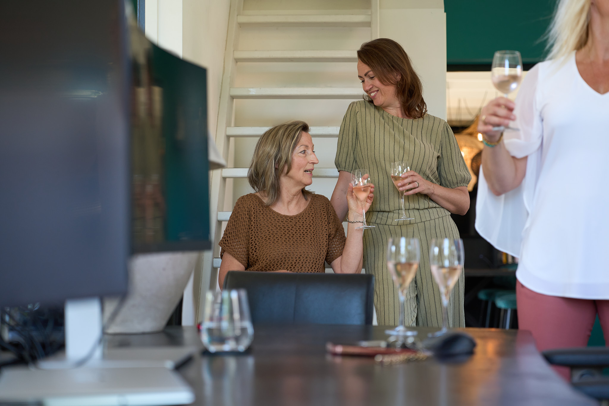 Two women toast with wine glasses on a white staircase, chatting and smiling, with a third guest partially visible in the foreground holding a glass.