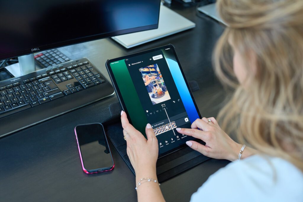 Person holds a tablet showing a video editing app while working at a desk with a keyboard and monitor nearby.