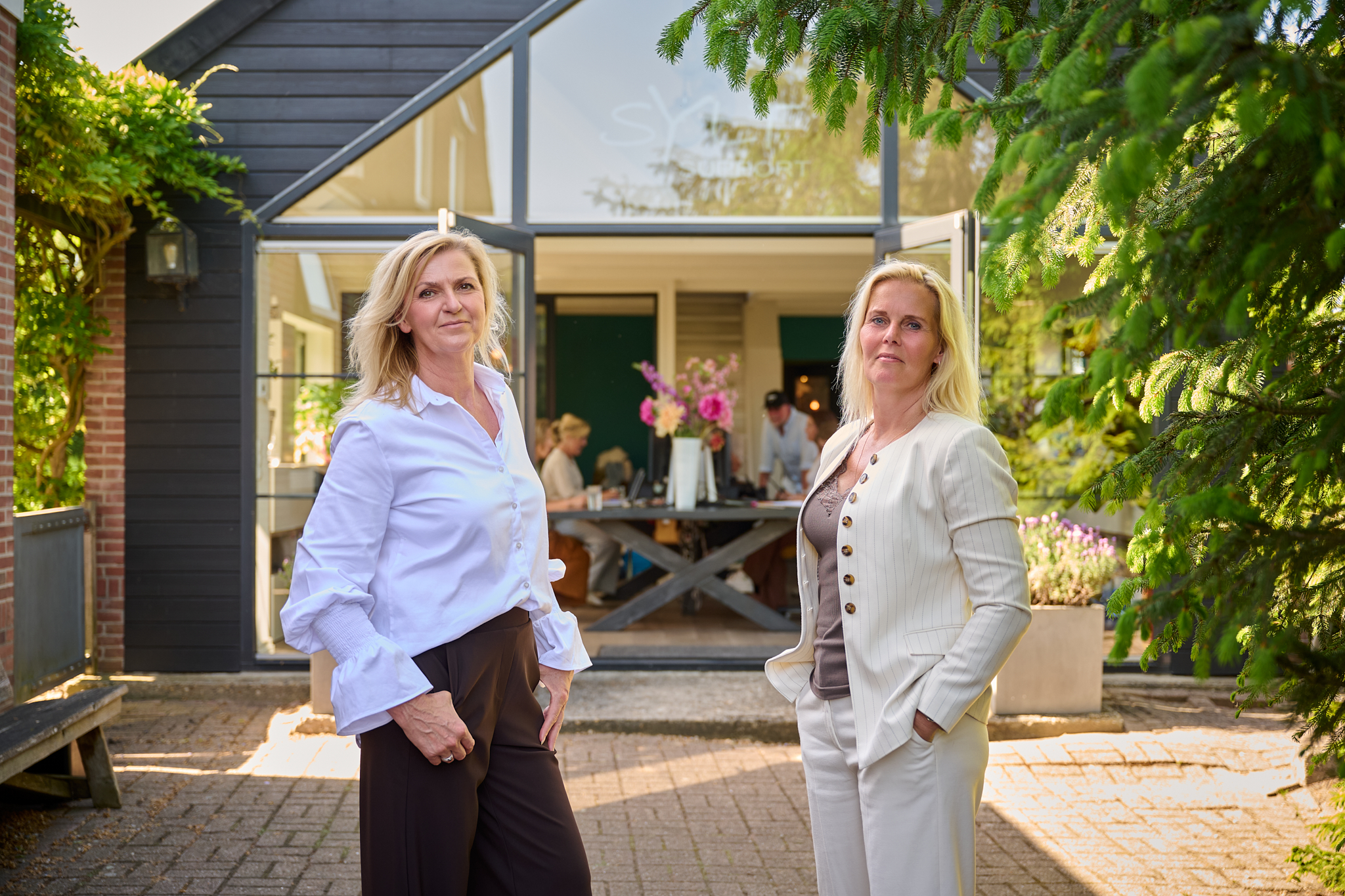 Two women in business-casual outfits stand outside a glass-front building, smiling, with greenery around.