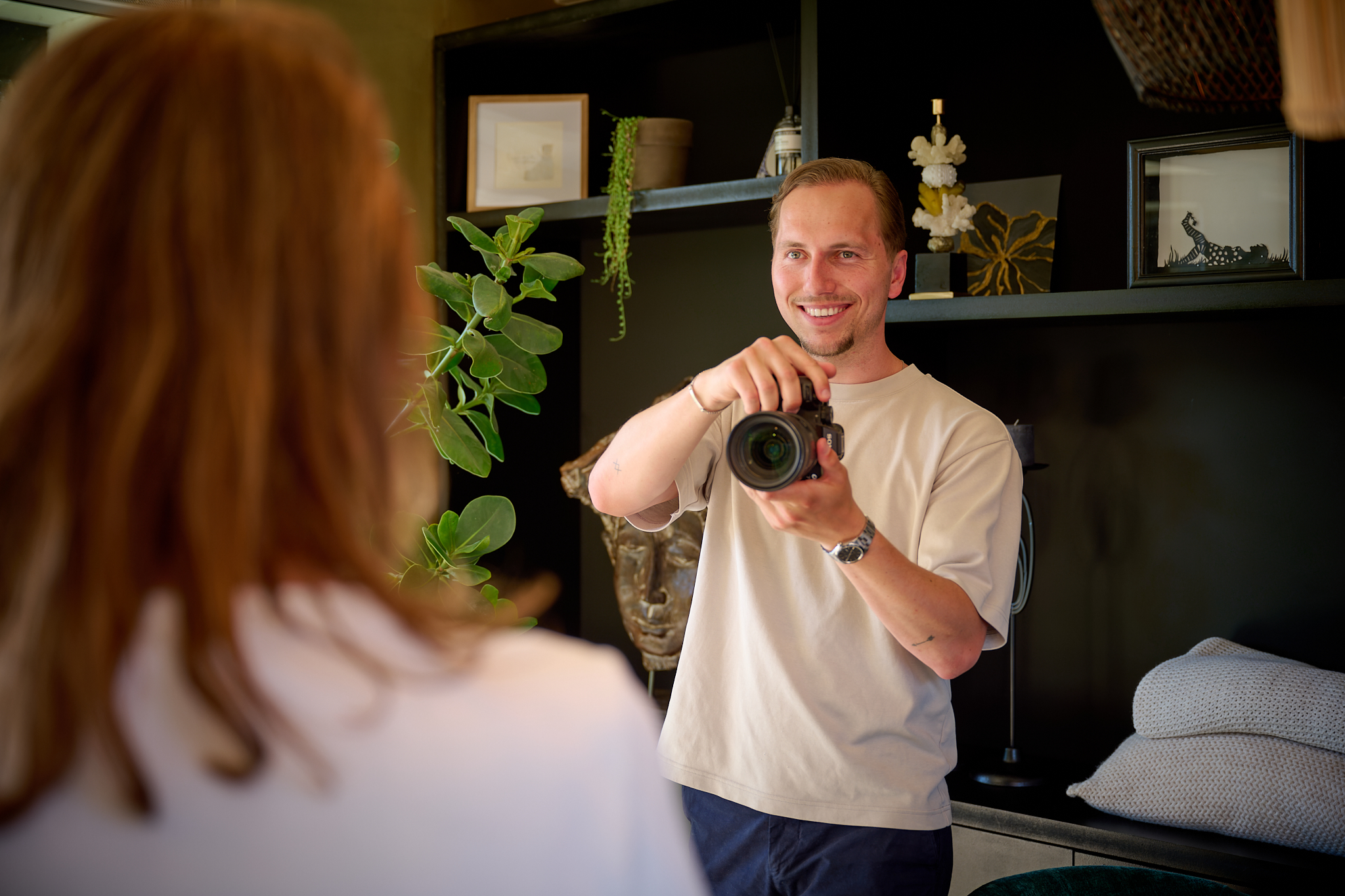 A man with a DSLR camera smiles as he photographs a person whose back is to the camera in a cozy living room with plants and shelves behind him.