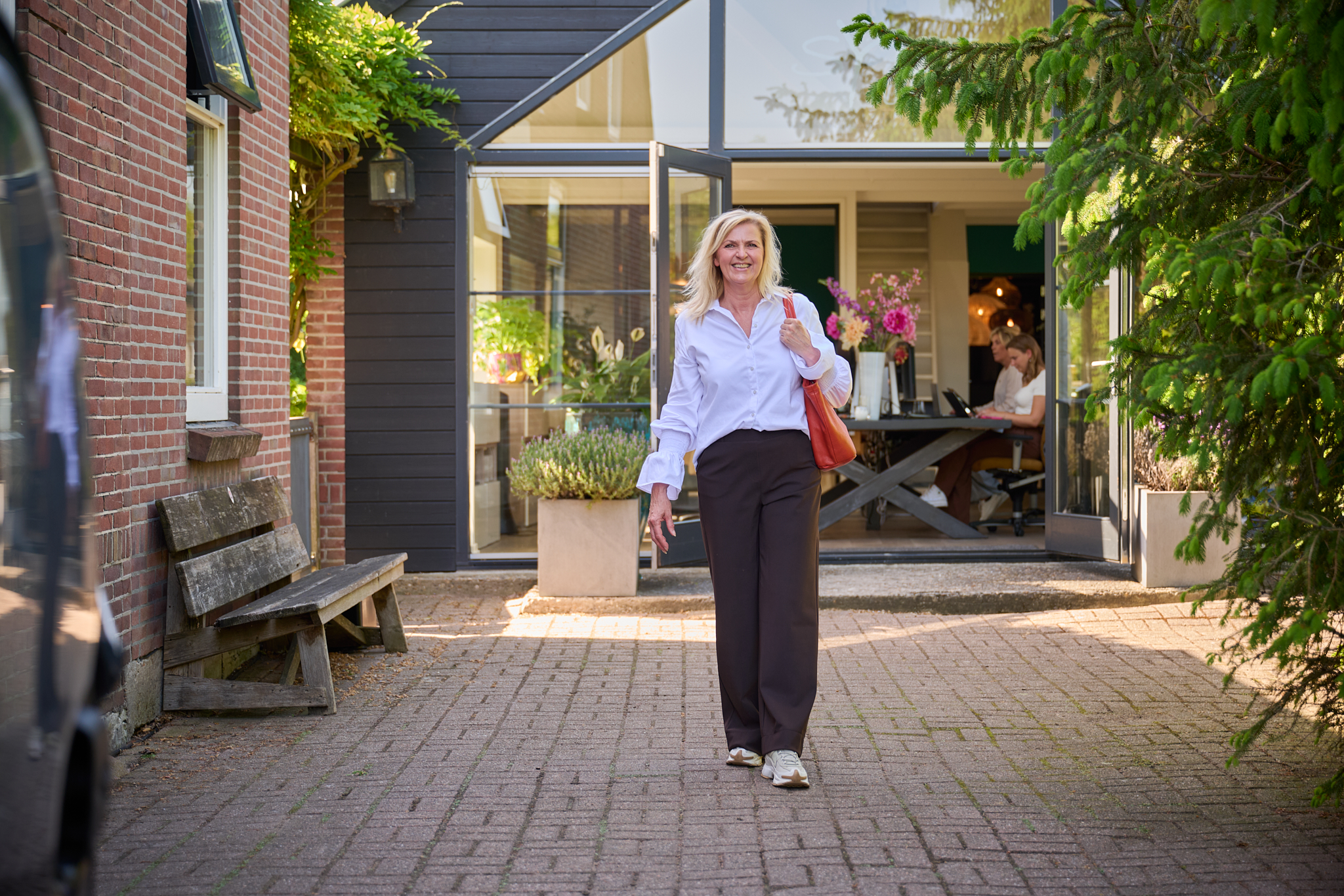 Smiling woman in a white blouse and dark trousers exits a glass-walled building, carrying an orange shoulder bag, with greenery around the doorway.