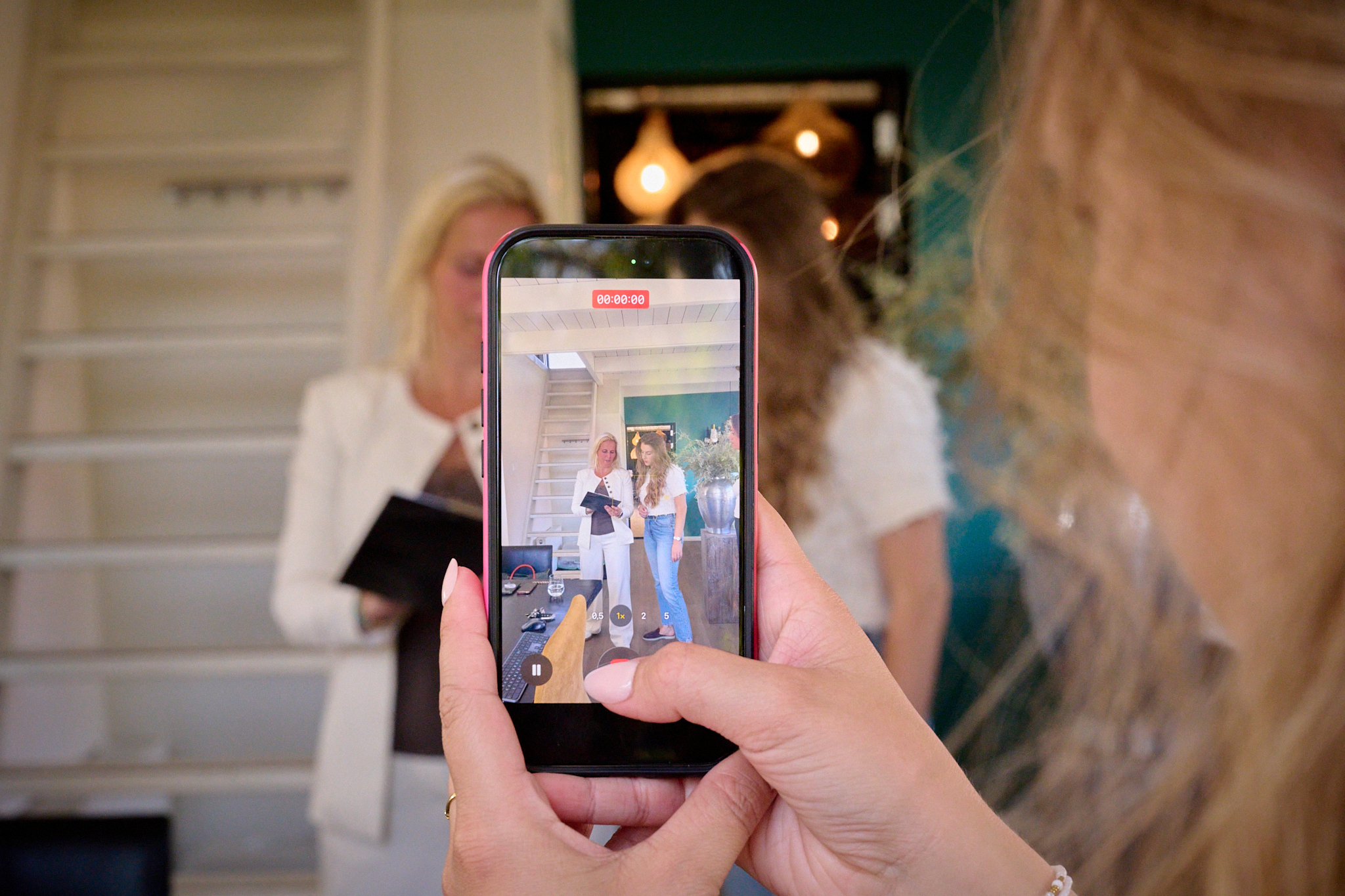 Hand holding a smartphone filming two women in a bright showroom with stairs in the background.