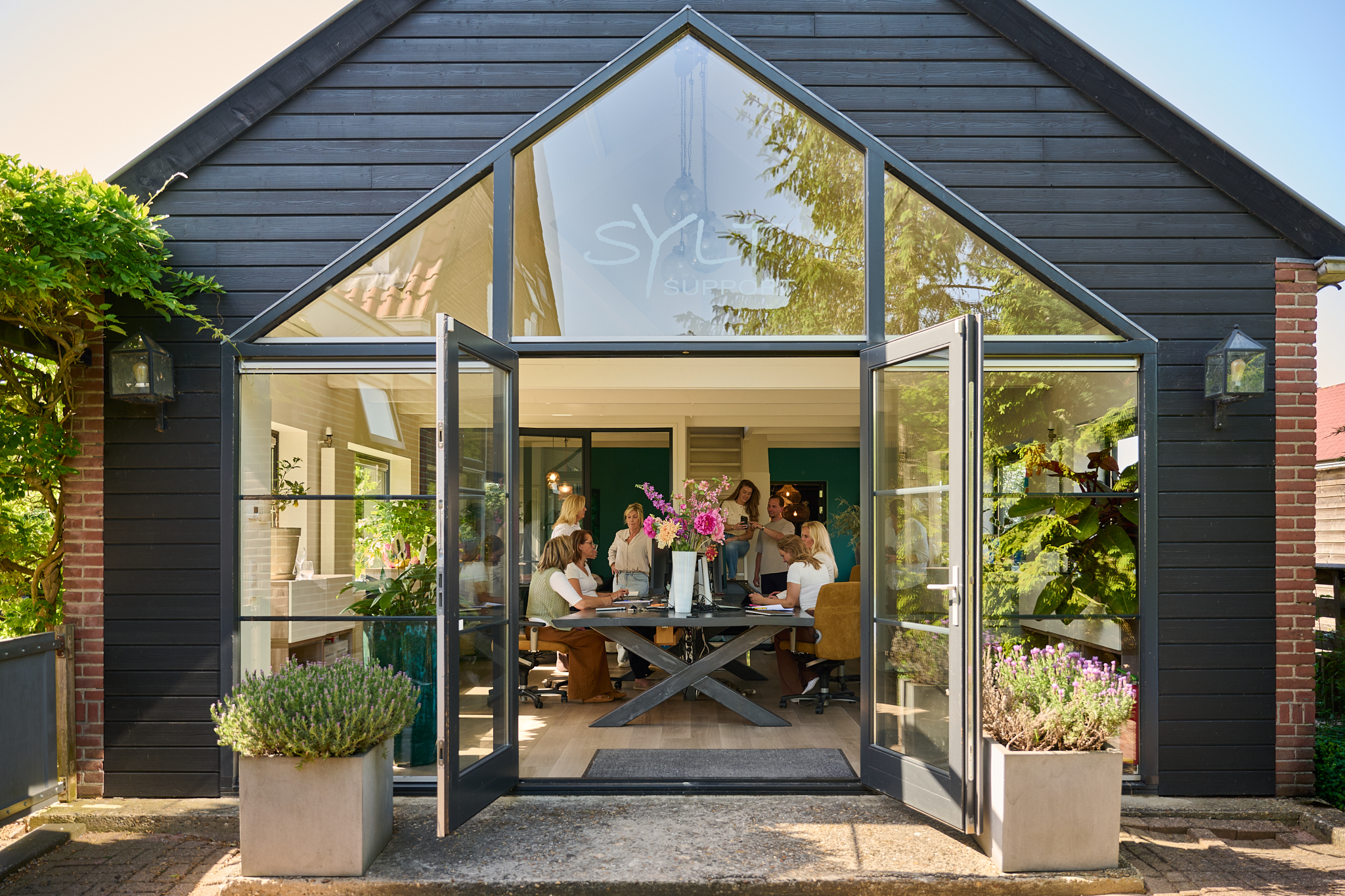 Group of people working together around a long table in a bright glass-walled meeting room, seen from outside through open double doors with plants nearby.