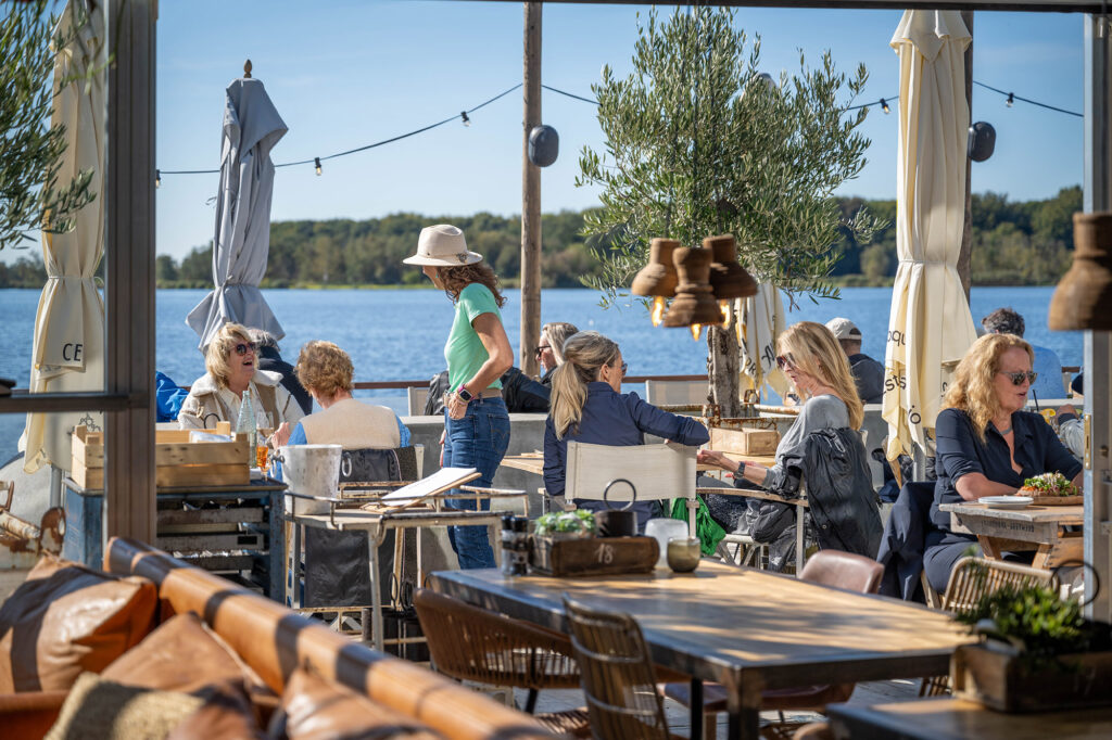 Group of people dining at an outdoor lakeside café with beige umbrellas and string lights over the tables by the water.
