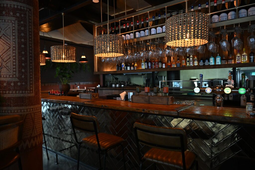 Cozy bar interior with hanging pendant lamps, bottles on shelves, and glassware above a wooden counter.
