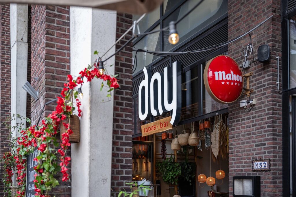 Brick storefront of a tapas bar with a round red Mahou sign and warm hanging lights. Temporarily visible ivy and red flowers frame the entrance.