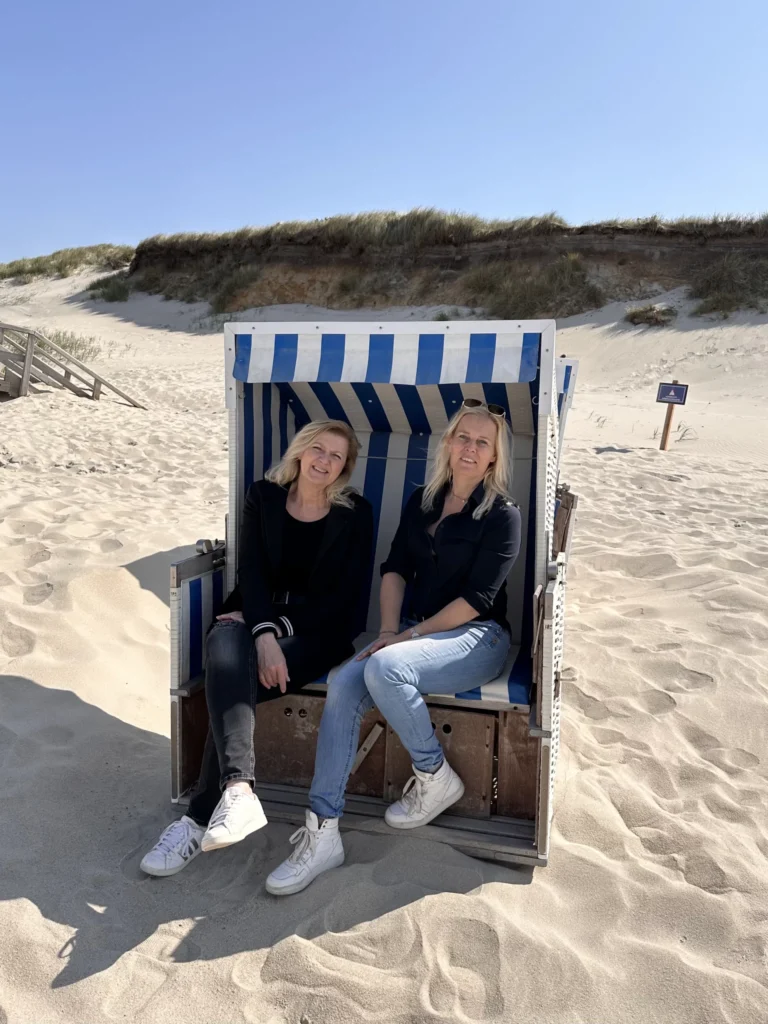 Two women sit inside a blue-and-white striped beach cabana on a sandy dune under a clear blue sky.