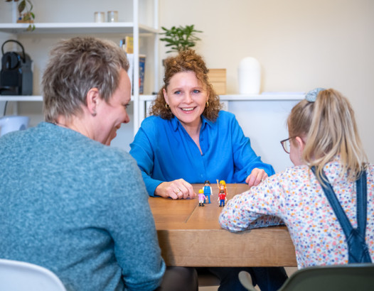 Three people sit around a wooden table, smiling as a woman in a blue shirt helps a young girl arrange small toy figures nearby a man watching.